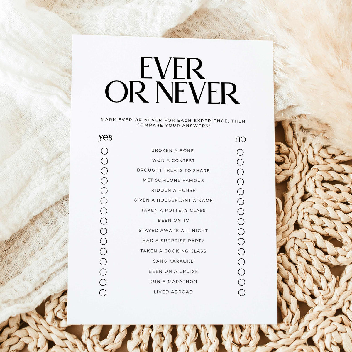 A white rectangular checklist card titled 'EVER OR NEVER' in black serif text, laid flat on a soft cream fabric with a beige woven rug underneath. The card lists experiences to mark ever or never, with circles for each item.