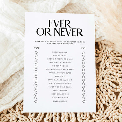 A white rectangular checklist card titled 'EVER OR NEVER' in black serif text, laid flat on a soft cream fabric with a beige woven rug underneath. The card lists experiences to mark ever or never, with circles for each item.