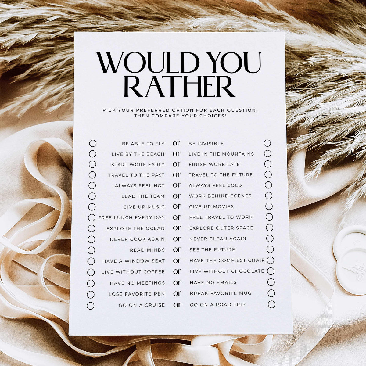 A white printable card with the large black text 'WOULD YOU RATHER' and a checklist of preference questions, styled on a beige fabric background with satin ribbons.