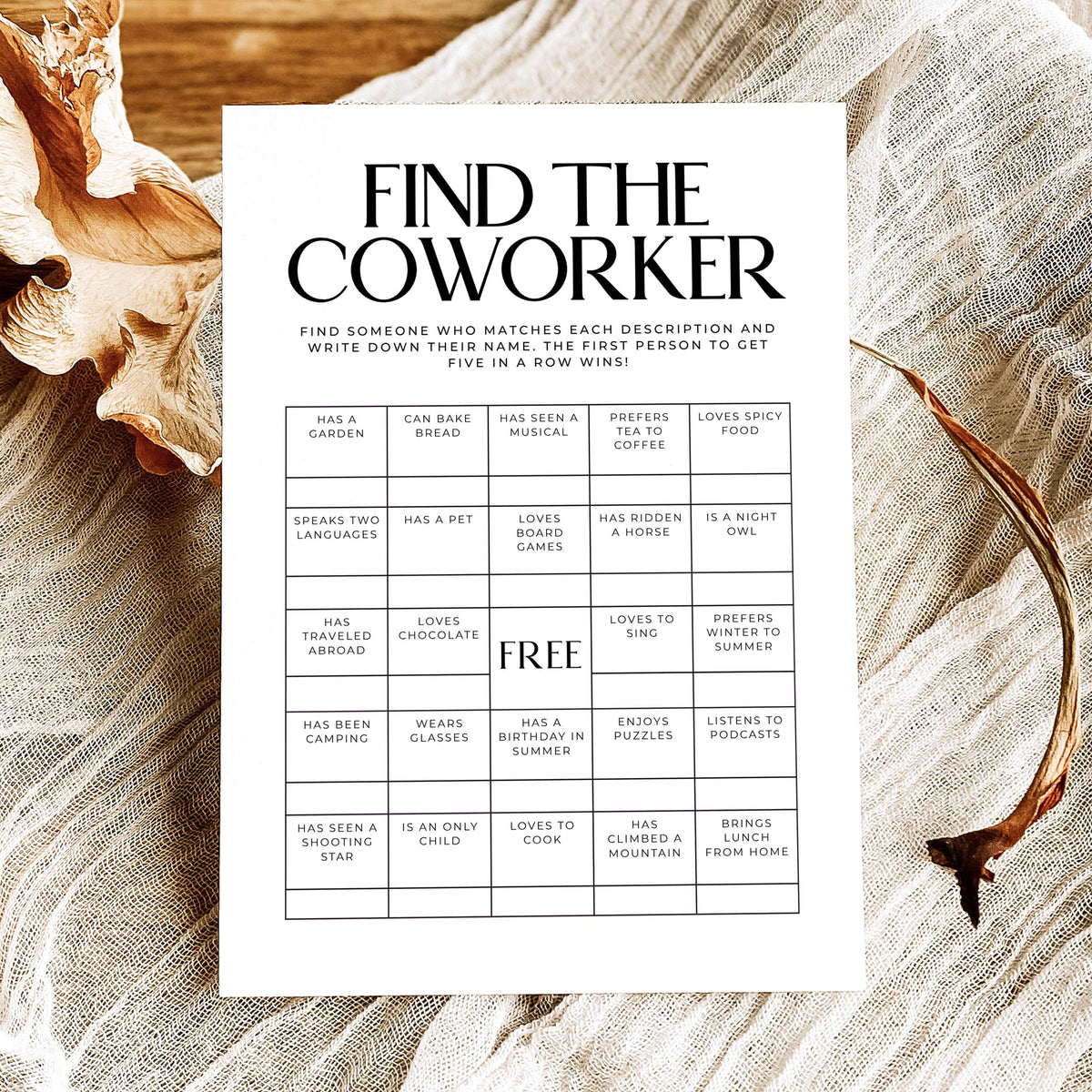 A printed 'Find the Coworker' game card with black text and a grid of clues, styled on a rustic wooden surface with linen fabric, dried flowers, and a twig.