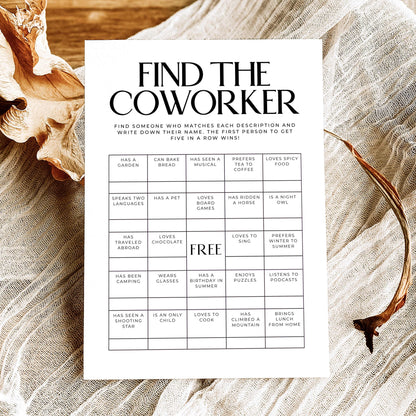 A printed 'Find the Coworker' game card with black text and a grid of clues, styled on a rustic wooden surface with linen fabric, dried flowers, and a twig.