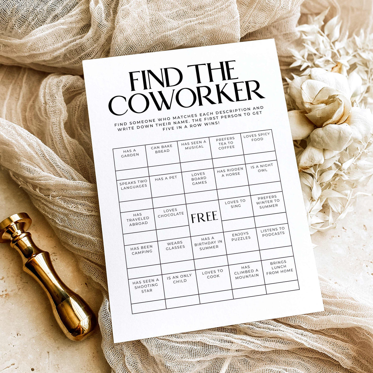 A printed 'Find the Coworker' activity card laid on beige fabric with dried flowers and a gold stamp, styled as a flat lay.