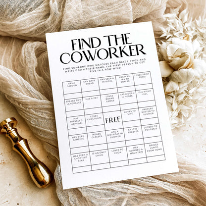 A printed 'Find the Coworker' activity card laid on beige fabric with dried flowers and a gold stamp, styled as a flat lay.