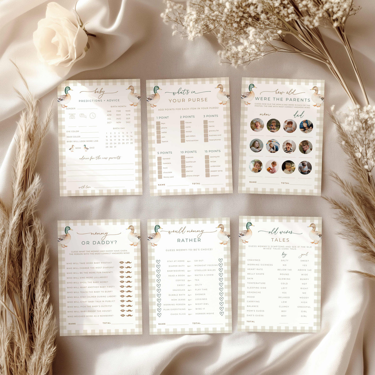 Six baby shower game cards arranged in two rows on a draped satin fabric backdrop with dried pampas grass and a white rose. The cards feature a soft beige and white gingham border, delicate teal duck illustrations, and various game prompts and photo-collage sections.
