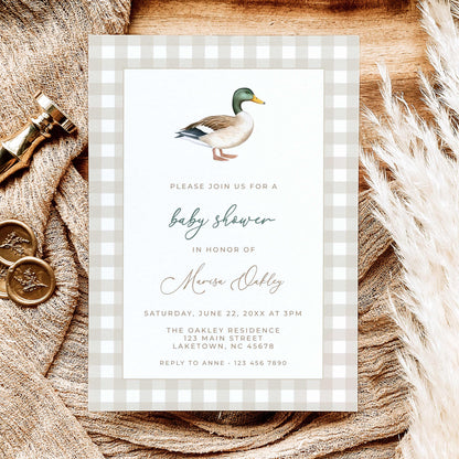 Mallard duck baby shower invitation featuring a watercolor mallard illustration, beige gingham border, and sage green script, displayed on natural linen with dried leaves.