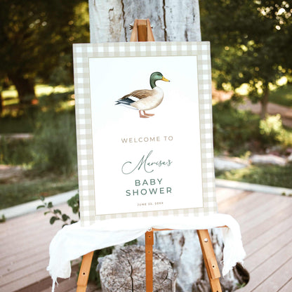 Mallard duck baby shower welcome sign featuring a watercolor mallard illustration, beige gingham border, and sage green lettering, displayed on a wooden easel outdoors.