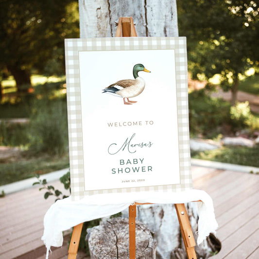 Mallard duck baby shower welcome sign featuring a watercolor mallard illustration, beige gingham border, and sage green lettering, displayed on a wooden easel outdoors.