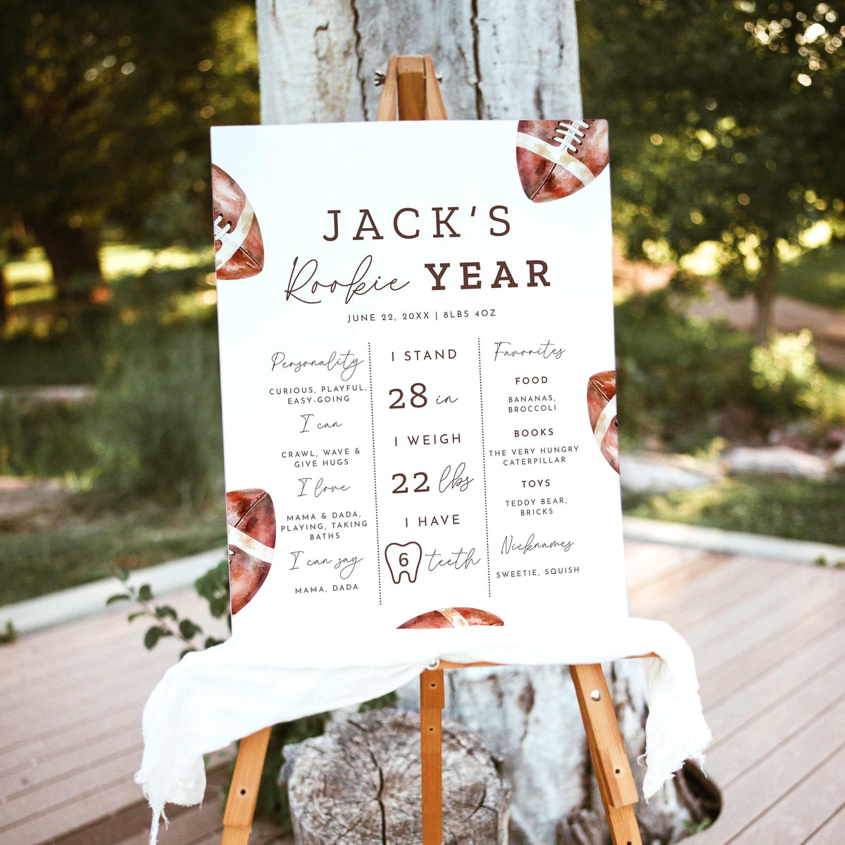 Rookie of the Year football first birthday milestone board featuring watercolor brown footballs, baby's height, weight, favorite things and first-year achievements, displayed on a wooden easel outdoors.