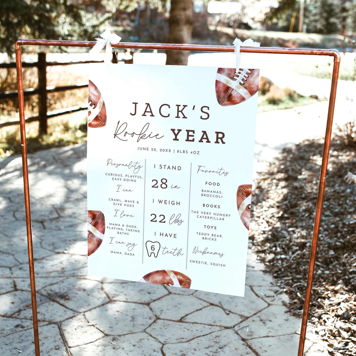 Rookie of the Year football first birthday milestone board featuring watercolor brown footballs, baby's height, weight, favorite things and first-year achievements, displayed on a wooden easel outdoors.