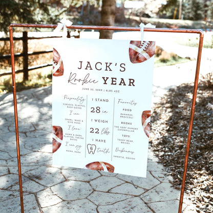Rookie of the Year football first birthday milestone board featuring watercolor brown footballs, baby's height, weight, favorite things and first-year achievements, displayed on a wooden easel outdoors.