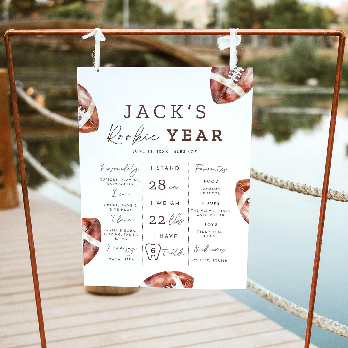 Rookie of the Year football first birthday milestone board featuring watercolor brown footballs, baby's height, weight, favorite things and first-year achievements, displayed on a wooden easel outdoors.