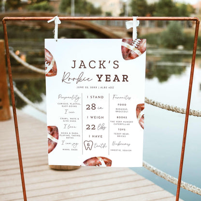 Rookie of the Year football first birthday milestone board featuring watercolor brown footballs, baby's height, weight, favorite things and first-year achievements, displayed on a wooden easel outdoors.